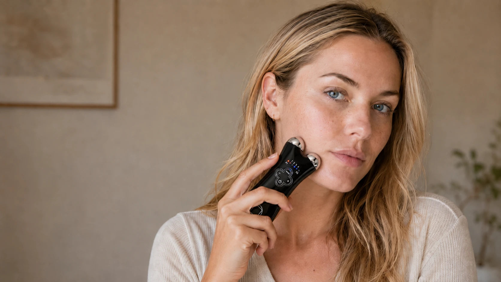 A woman in her thirties using the black-and-chrome NRGY microcurrent device along her cheek at home, representing Truth Treatment Systems exfoliate and energize collection